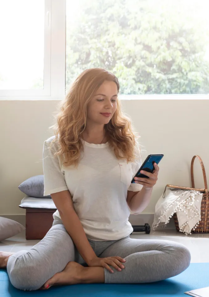 Woman seated on a yoga mat checking her phone by a sunny window